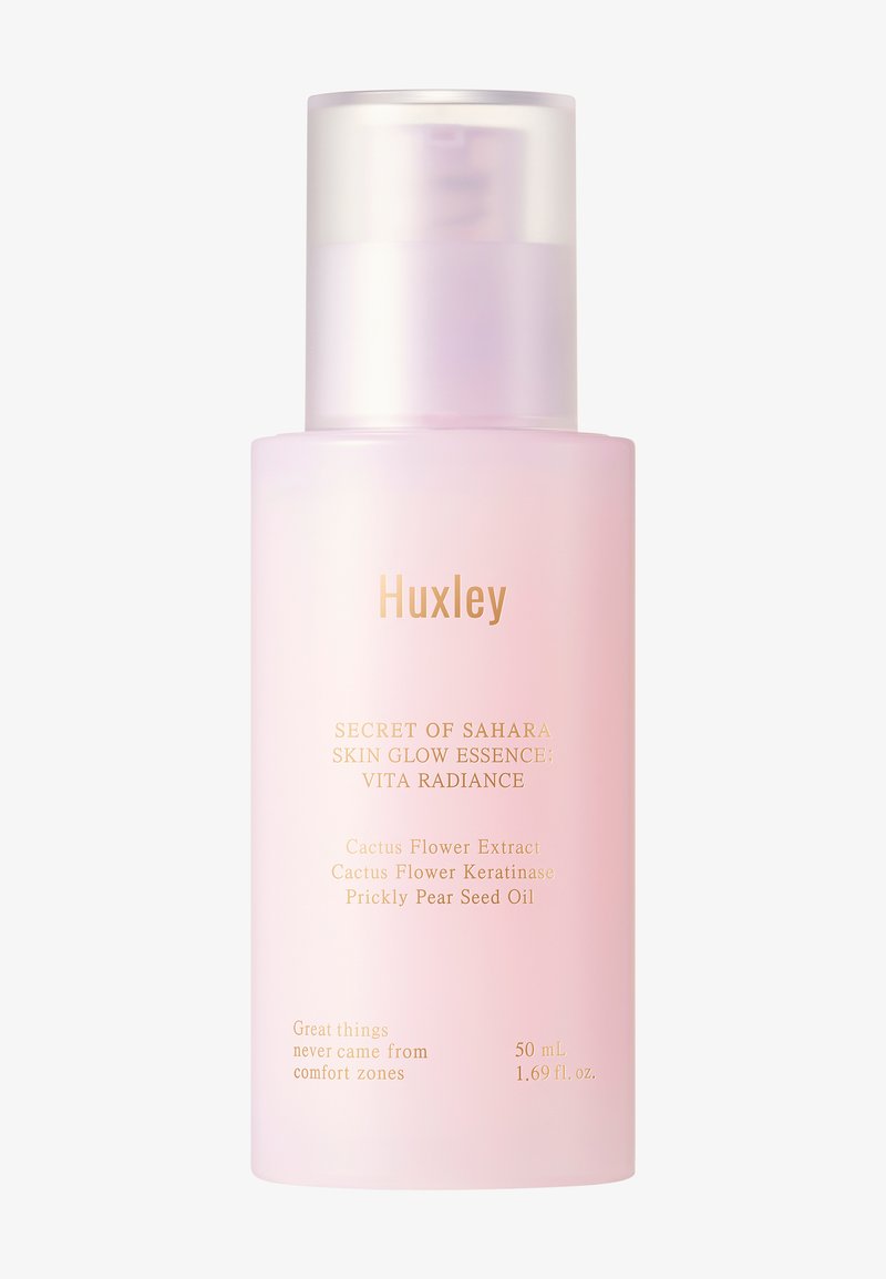 Huxley Skin Glow Essence in een doorschijnende roze fles met gouden tekst. Beschikt over een pompdispenser; bevat cactusbloem- en prickly pear-extracten.