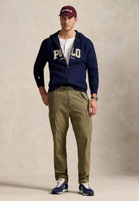 Camisola com fecho éclair azul-marinho com logótipo "POLO", camisa henley branca, calças cargo verde-oliva, cinto verde e sapatilhas azul-marinho. Boné bordô com texto "NEW ENGLAND".