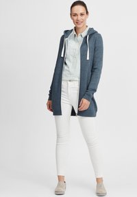 Mörkblå zip-up hoodie i mjukt material, med huva, vita snoddar och slim fit, kombinerad med vita smala byxor och ljusa skor.