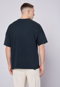 Per didelė tamsiai mėlyna medvilninė marškinėliai trumpomis rankovėmis. Teksūruotas vientisas audinys ir apvalus kaklas, derinami su šviesiomis kelnėmis.