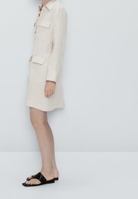 Beiges Leinenkleid mit Kragen, Schnürung vorne, zwei Brusttaschen und langen Ärmeln. Kombiniert mit schwarzen flachen Sandalen.