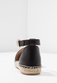 Sandal de cuero negro con parte trasera redondeada, tirante en el tobillo y hebilla dorada, que cuenta con una suela de yute trenzado y una textura suave.