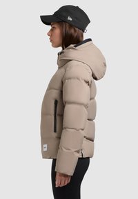 Chaqueta de plumas beige con capucha, que presenta herrajes y cremalleras negras. Una gorra negra completa el look, mostrando un diseño elegante y moderno.