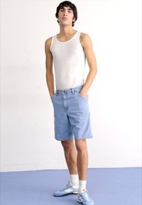 Top blanco sin mangas, pantalones cortos de mezclilla azul y zapatillas azul claras con detalles blancos. La modelo está de pie con las manos en los bolsillos, en un fondo neutro.