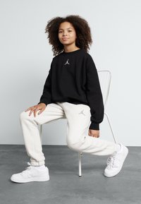 Sudadera negra con un pequeño logotipo plateado, combinada con pantalones de chándal gris claro que presentan un logotipo similar. Zapatillas deportivas blancas completan el atuendo.