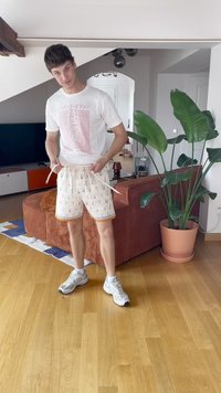 Mladý muž uvnitř, který zavazuje šňůrku na světlých barevných šortkách s potiskem, má na sobě bílý tričko s grafikou a tenisky, v pozadí je rostlina a pohovka.