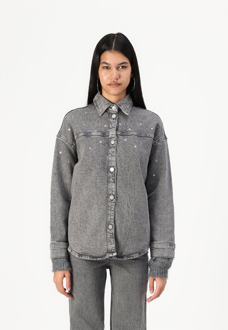 Veste en denim grise avec un devant à boutons, un col pointu et des clous argentés. Elle présente des manches longues et une coupe décontractée.