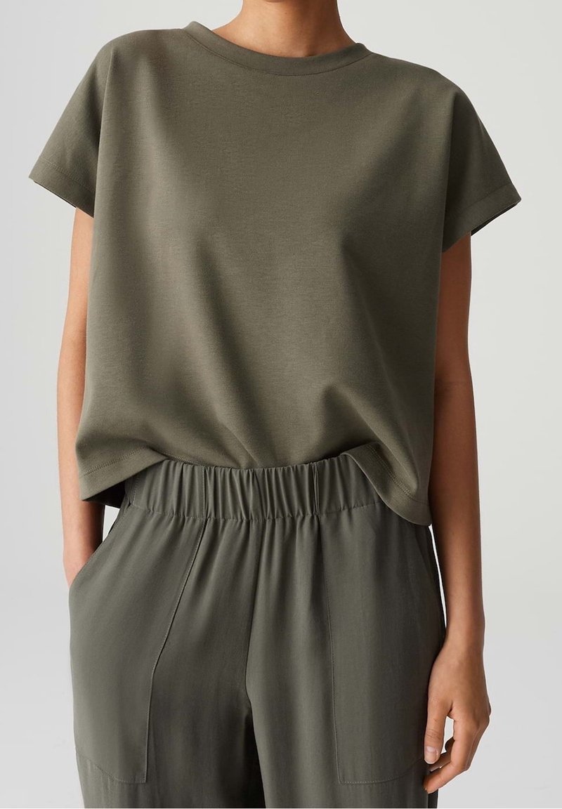 Personne portant un haut olive à manches courtes ample, légèrement rentré dans un pantalon taille haute olive avec taille élastique et poches latérales.