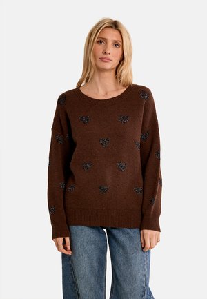 Donna con capelli biondi che indossa un maglione marrone con motivi a cuore neri e jeans blu su uno sfondo chiaro uniforme.
