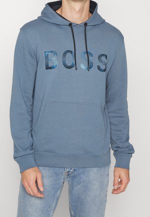 Homme portant un sweat à capuche bleu avec poche avant et logo rayé brillant "BOSS", associé à un jean bleu clair, debout devant un fond uni.