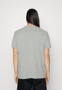 Persona con lunghi capelli scuri che indossa una semplice t-shirt grigia a maniche corte e pantaloni neri, vista di spalle su uno sfondo bianco.