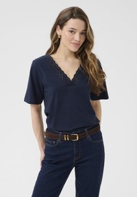Marineblå T-shirt med V-hals og bølgemønster af blondekant. Korte ærmer, parret med mørke denimjeans og et brunt bæltet.