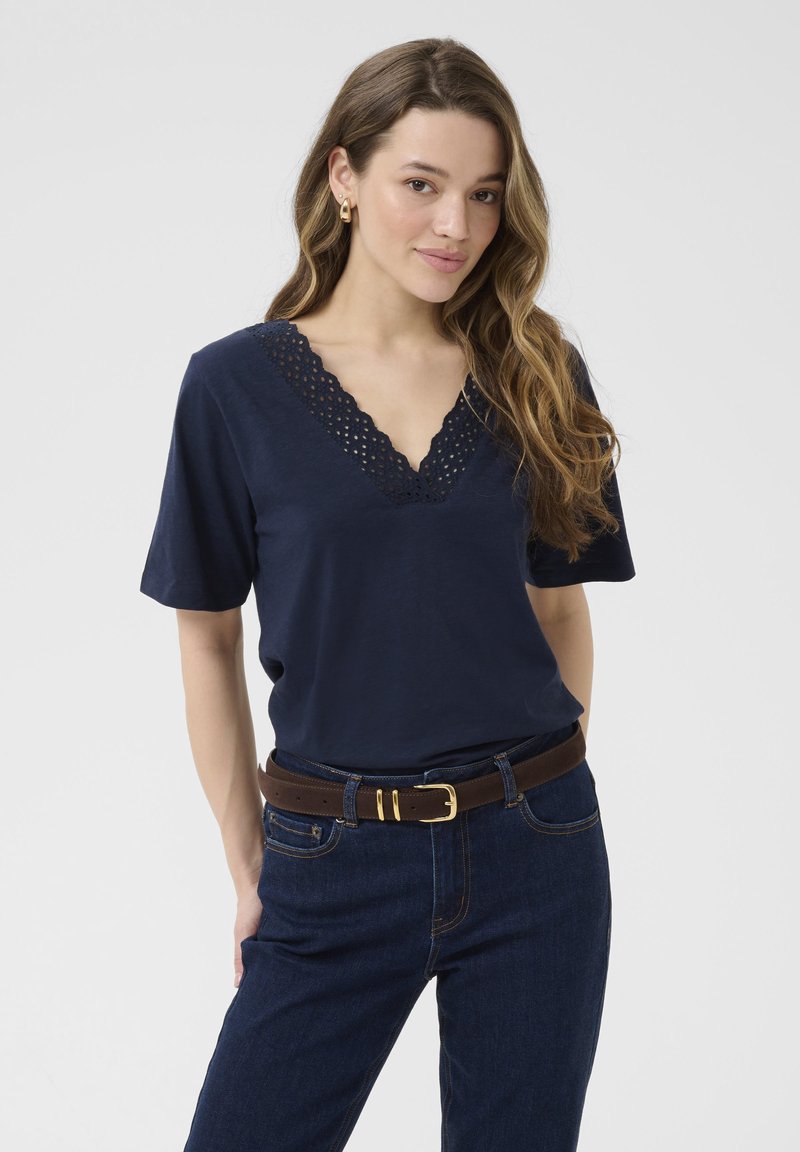 Marineblå T-shirt med V-hals og bølgemønster af blondekant. Korte ærmer, parret med mørke denimjeans og et brunt bæltet.