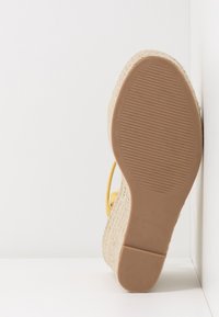 Semelle d'une sandale compensée beige avec texture tissée et semelle inférieure en caoutchouc marron, positionnée verticalement contre une surface blanche.