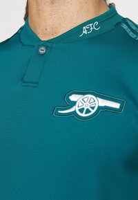 Polo bleu sarcelle avec un col boutonné, présentant un logo canon blanc brodé et le texte "AFC" sur le col, fabriqué en tissu lisse.