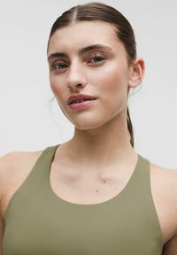 Giovane donna con pelle chiara e capelli scuri raccolti, indossa una canotta verde oliva, guarda leggermente di lato su uno sfondo neutro.