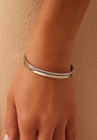 Pulsera de metal plateado con un diseño elegante y curvado. Un lado presenta una fila de piedras claras y brillantes, ofreciendo un contraste sutil en textura.