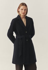 Marineblauer Wollmantel mit tailliertem Schnitt, Reverskragen und zwei vorderen Knöpfen. Glatte Textur und maßgeschneiderte Passform, kombiniert mit braunen Hosen.