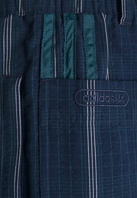 Mörkblå rutiga byxor med vertikala vita och gröna ränder, elastisk midja och en broderad logotyp. Har fickor och banddetaljer.