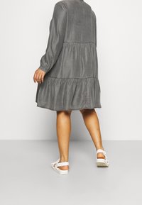 Grijze gelaagde jurk met lange mouwen en ronde halslijn, gemaakt van lichte stof. Witte sandalen met banden worden gedragen aan de voeten.