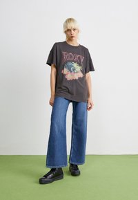 T-shirt graphique gris avec le logo "ROXY" et une scène de plage ; associé à un jean bleu à jambes larges et des chaussures noires à lacets.