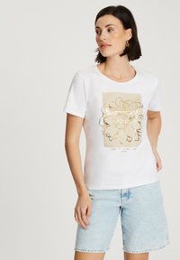 Biała koszulka z krótkim rękawem z złotym graficznym wzorem kwiatowym na jasnobeżowym tle. Miękka tkanina, okrągły dekolt i luźny krój.
