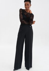 Kobieta ubrana w czarną, przezroczystą bluzkę w kwiaty z długim rękawem, czarne, wysokie szerokie spodnie i kolczyki w kształcie kół, stojąca na białym tle.