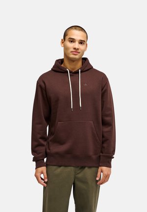 Jeune homme portant un sweat à capuche marron avec des cordons blancs et un pantalon vert olive, debout devant un fond clair uni.