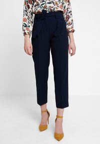 Pantalon bleu marine cintré avec taille haute, plis sur le devant et ceinture en tissu. Assorti à un chemisier à manches longues à imprimé floral et des escarpins à bride cheville moutarde.