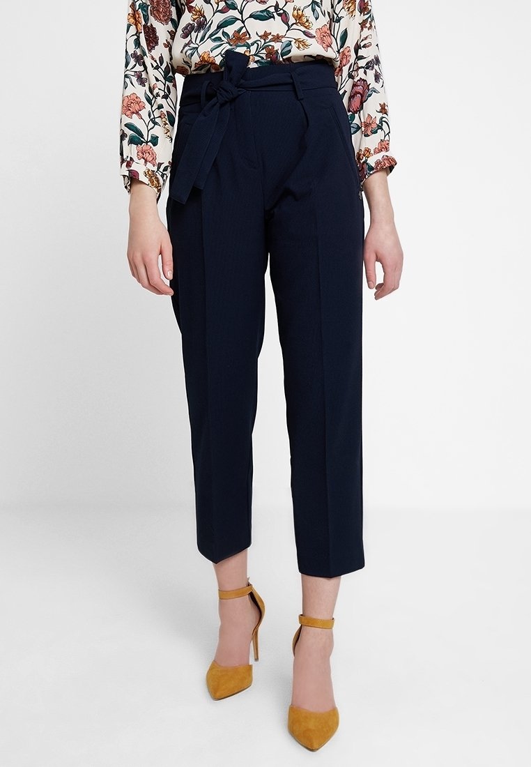 Pantalon bleu marine cintré avec taille haute, plis sur le devant et ceinture en tissu. Assorti à un chemisier à manches longues à imprimé floral et des escarpins à bride cheville moutarde.