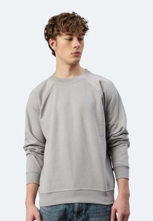 Giovane uomo con capelli ricci che indossa una felpa grigio chiaro a maniche lunghe e jeans blu, guarda leggermente verso il basso a sinistra.