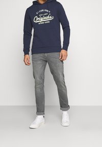 Tmavě modrá mikina s bílým logem, bavlněný materiál, dlouhé rukávy, přední kapsa, kombinována se světle šedými džíny a bílými teniskami.