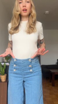 Γυναίκα με μακριά ξανθά μαλλιά, που φοράει λευκό πουκάμισο και ψηλόμεσο μπλε τζιν παντελόνι, κάνει χειρονομίες με τα χέρια σε ένα σαλόνι με φυτά και έπιπλα.