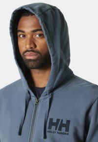 Sudadera azul con cremallera hecha de un material suave, con capucha, cremallera en la parte delantera y un logotipo negro en la parte izquierda del pecho.