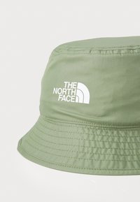 Lichtgroene bucket hat van textuurstof. Heeft een wit logo aan de zijkant en gestikte details langs de rand.