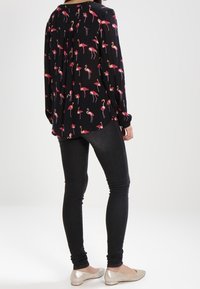 Schwarze Bluse mit langen Ärmeln und Flamingo-Print, kombiniert mit schwarzen Skinny Jeans und metallischen Flats. Weicher Stoff mit lockerer Passform.