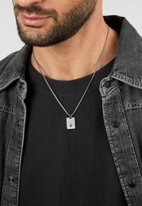 Collana con pendente d'argento su una catena grigia texture, con un ciondolo rettangolare decorato con un design a stella, indossata su una maglietta e una giacca nera.
