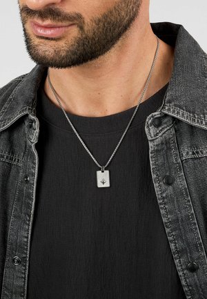 Collana con pendente d'argento su una catena grigia texture, con un ciondolo rettangolare decorato con un design a stella, indossata su una maglietta e una giacca nera.
