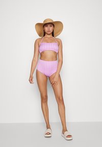 Conjunto de biquíni com padrão de gingham rosa, composto por um top curto e uma parte de baixo de cintura alta. Acompanhado de um chapéu de palha de aba larga e sandálias de slide em tons neutros.