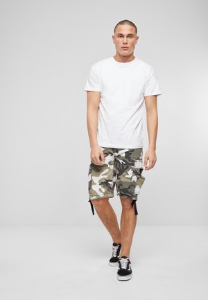 T-shirt bianco in cotone abbinato a pantaloni cargo verdi mimetici con molte tasche e sneaker nere con lacci. Sfondo semplice.