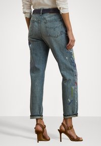 Gaiši zili džinsi ar brīvu piegriezumu, ar daudzkrāsainu krāsas šļakatu detaļām un sarullētām maliņām. Apvienoti ar brūniem augstpapēžu sandalēm.