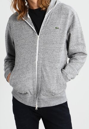 Sudadera con cremallera gris hecha de tela texturizada, con una cremallera blanca y un logo verde en el lado izquierdo del pecho, con bolsillos frontales y capucha.