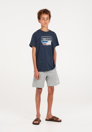 Marineblaue Baumwoll-T-Shirt mit bedrucktem "PROTEST"-Logo, kombiniert mit grauen Shorts und braunen Sandalen. Einfaches Design, lockere Passform.