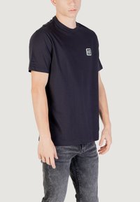 T-shirt in cotone blu navy con collo a giro e maniche corte. Presenta un piccolo logo quadrato sul petto. Indossato con jeans grigi scuri.