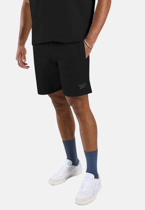 Hombre vistiendo pantalones cortos negros de Reebok, camiseta negra, calcetines azules y zapatillas blancas, de pie con una mano en el bolsillo contra un fondo blanco.