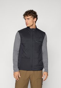 Zwarte gewatteerde vest met een hoge kraag, rits aan de voorkant en een borstzak. Grijze langlopende shirt eronder en beige broek.