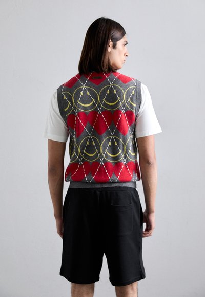 Gestrickte Weste in Rot, Grau und Gelb mit Muster aus Rauten. Kombiniert mit schwarzen Shorts, die über einen elastischen Bund und eine Seitentasche verfügen.