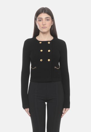 Donna che indossa un cardigan nero aderente con bottoni dorati e dettagli a catena, abbinato a pantaloni neri a vita alta, in piedi su sfondo bianco.