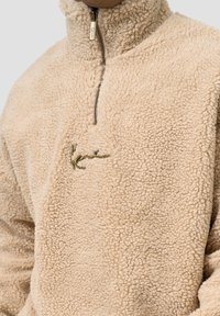 Beige fleece-genser med høy krage, halv glidelås og brodert grønn logo. Stoffet har en myk, strukturert overflate.