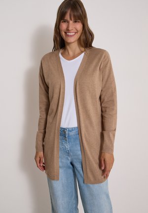 Smilende kvinde iført en beige åben cardigan over en hvid skjorte og lyseblå højttaljede jeans, stående mod en ensfarvet baggrund.